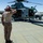 A U.S. Marine in desert camouflage utilities renders a salute to a military helicopter numbered '90' on a desert airfield tarmac. The helicopter, identifiable as a Bell UH-1Y Venom based on its profile, has its rotors spinning and crew members visible inside. The arid, desert environment and 'MARINES' markings on the aircraft suggest this is likely a U.S. Marine Corps installation in a southwestern desert region.