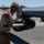 A U.S. Marine in desert MARPAT camouflage uniform is loading or inspecting munitions on an aircraft wing pylon on a tarmac. The setting appears to be a desert airbase, with arid terrain and mountains visible in the background. The activity suggests pre-flight ordnance loading or maintenance operations.