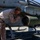 A U.S. Marine in desert MARPAT camouflage uniform is performing maintenance or inspection work on a rocket pod mounted to a military helicopter on a tarmac. The rocket pod appears to be a 7-tube or 19-tube unguided rocket launcher (likely an M260 or M261 HYDRA 70 system) attached to the skid-mounted hardpoint of what appears to be a Bell AH-1Z Viper or similar attack helicopter. The setting appears to be a desert airfield under clear, sunny conditions.