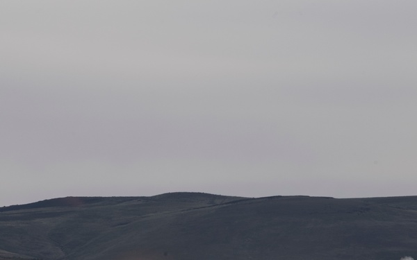 A rocket or missile is being launched from a ground-based launcher system in an arid, open terrain environment. A large plume of white smoke and dust billows from the launch site, while the projectile is visible in flight with a bright flame trail. The setting appears to be a military test range or training exercise conducted at dusk or under overcast conditions.