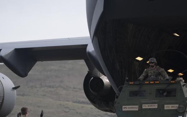 A military loading operation is underway at an airfield, where a HIMARS (High Mobility Artillery Rocket System) vehicle is being loaded into or unloaded from a large military transport aircraft, likely a C-17 Globemaster III. A ground guide in camouflage uniform is directing the vehicle operator using hand signals. The setting appears to be a semi-arid region with rolling hills visible in the background.