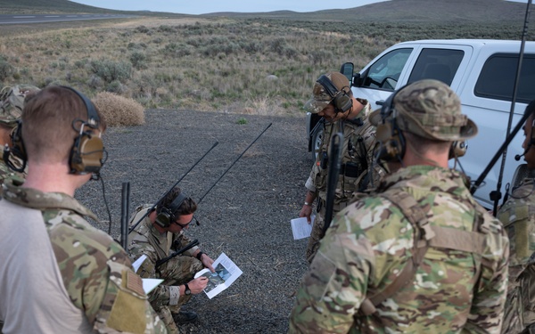 A group of military personnel in MultiCam/OCP camouflage uniforms are gathered in a semi-circle on a gravel surface in an arid, open landscape. They appear to be conducting a briefing or mission planning session, reviewing printed documents and photographs. Several personnel are equipped with tactical headsets and radio antennas, suggesting active communications.