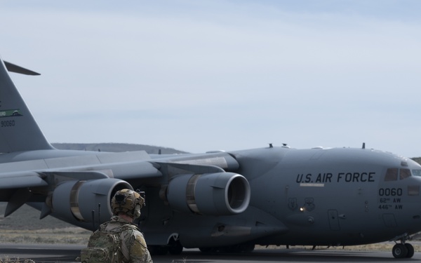A U.S. Air Force C-17 Globemaster III military transport aircraft is taxiing on a runway in an arid, semi-desert environment. A soldier in full combat gear and helmet is seated on a small dirt/trail motorcycle in the foreground, observing the aircraft. The aircraft markings indicate it belongs to the 62nd Airlift Wing and 445th Airlift Wing, with tail number 0060.
