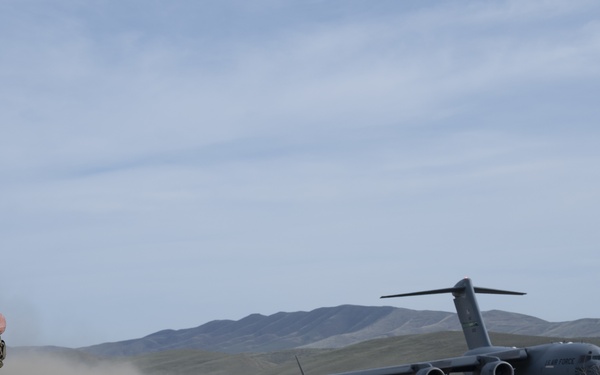 A military personnel in camouflage uniform stands on a gravel airstrip holding a radio, observing a U.S. Air Force C-17 Globemaster III transport aircraft on the runway. The aircraft is either landing or taking off on what appears to be an austere or forward operating airstrip in a semi-arid, hilly terrain. Dust is being kicked up behind the aircraft, suggesting it is operating on an unpaved or semi-prepared surface.