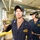 A U.S. Navy sailor in dark blue coveralls holds up a military patch for the USS Monsoon (PC 4), assigned to LT Mitchell Kempisty, OPS/NAV. Both personnel are wearing communication headsets and dark navy coveralls with unit patches, standing in what appears to be an industrial or shipboard interior environment. A second sailor stands in the background, also wearing a headset.