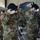 A group of U.S. Army soldiers in OCP (Operational Camouflage Pattern) uniforms are simultaneously donning dark-colored berets during what appears to be a formal military ceremony held inside an aircraft hangar. Several soldiers are wearing face masks, suggesting this event took place during the COVID-19 pandemic period. White folding chairs and a military aircraft are visible in the background.
