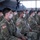 A large group of U.S. Army soldiers in OCP (Operational Camouflage Pattern) uniforms are seated in rows of white folding chairs inside a military hangar. All personnel are wearing white face masks, consistent with COVID-19 era health protocols. A military aircraft, likely a C-130 Hercules, is visible in the background.