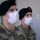 Two U.S. Army soldiers wearing green berets and white surgical face masks are shown in a military setting. The soldier in the foreground is in sharp focus and displays a 'U.S. ARMY' tape on their OCP (Operational Camouflage Pattern) uniform, while the second soldier is blurred in the background. Both wear the Special Forces green beret, with the in-focus soldier's beret featuring a red flash with the Special Forces crest.