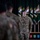 A U.S. Army soldier wearing a green beret stands at attention during what appears to be a formal military ceremony. The soldier, identified by name tape as 'TOOHEY' and wearing the rank of Major (oak leaf cluster), is positioned in front of multiple Special Forces unit flags. Several other uniformed soldiers are visible in the blurred foreground.
