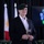 A middle-aged man wearing a dark green military beret and a black blazer over a white shirt stands holding a wine glass with red wine. He appears to be at a formal military or ceremonial event, evidenced by the flags and military insignia display visible in the background. His posture and attire suggest he may be a veteran or civilian dignitary attending an official function.