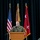 A U.S. Army officer is speaking at a podium during a formal military ceremony. The podium displays a shield emblem with the Latin motto 'VERITAS ET LIBERTAS' and what appears to be a unit crest. Three flags are visible behind the speaker, including the U.S. flag and a red general officer flag with two stars.