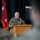A U.S. Army officer is speaking at a wooden podium during what appears to be a formal military ceremony or briefing. The speaker is in Army Combat Uniform (ACU/OCP pattern) and is addressing an audience of uniformed personnel. A red general officer flag with two stars is visible in the background, indicating a two-star (Major General) command presence.