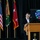 A civilian male speaker addresses an audience from a wooden podium at what appears to be a U.S. Army Special Forces official ceremony or event. Behind him are three flags including the U.S. flag, a green unit flag, and a two-star general officer flag. A screen displaying the U.S. Army Special Forces insignia and group patches is visible in the background.