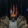A civilian speaker in formal attire addresses a large assembly of military personnel in camouflage uniforms at what appears to be an official military ceremony. The speaker stands at a wooden podium bearing a military crest/seal, flanked by three flags including the U.S. flag and a red general officer flag. Rows of seated soldiers in Army Combat Uniforms (ACU) face the podium in a formal indoor venue with black curtain backdrop.