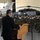 A civilian speaker in a dark suit addresses a large formation of military personnel seated in a hangar. The audience, wearing face masks, is seated in rows facing the podium. Military aircraft, including what appears to be a helicopter, are visible in the background of the hangar.
