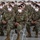A large formation of U.S. Army soldiers seated in rows on white folding chairs, all wearing white face masks and multicam/OCP camouflage uniforms. The setting appears to be an indoor facility such as a hangar or large assembly hall. The gathering suggests a formal briefing, ceremony, or official event during the COVID-19 pandemic era.