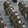 A large group of military personnel in U.S. Army camouflage uniforms (OCP pattern) are seated in socially distanced rows on white folding chairs inside what appears to be a hangar or large indoor facility. All individuals are wearing white face masks, consistent with COVID-19 pandemic health protocols. The formation suggests a briefing, ceremony, or official gathering.