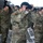 A group of U.S. Army soldiers in OCP (Operational Camouflage Pattern) uniforms are simultaneously removing or placing black berets on their heads during what appears to be a formal military ceremony held indoors. The soldiers are wearing face masks, suggesting this event took place during the COVID-19 pandemic period. Folding chairs are arranged in the background, indicating a structured ceremonial formation.
