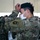 A U.S. Army Special Forces soldier is adjusting a green beret on his head during what appears to be a formal military ceremony. The soldier is wearing OCP (Operational Camouflage Pattern) uniform with a Special Forces Airborne patch visible on the left sleeve and a white face mask. In the background, rows of other uniformed soldiers wearing face masks are visible, suggesting a group ceremony held indoors.