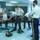 A group of military cadets in uniform are presenting and demonstrating a robotics project in a classroom setting. The project, displayed on a poster board, is titled 'Reinforcement Learning on Modular Robots using Real-Time Decentralized Deconfliction.' Several small wheeled robots are deployed on the floor as part of the live demonstration.