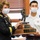 Two individuals wearing face masks are jointly holding a military trophy/award. The person on the left is wearing a U.S. Army dress uniform with the name tag 'JEBB,' while the person on the right appears to be a cadet in a white uniform. The trophy features a bronze soldier figurine on top of a wooden base with engraved plaques.