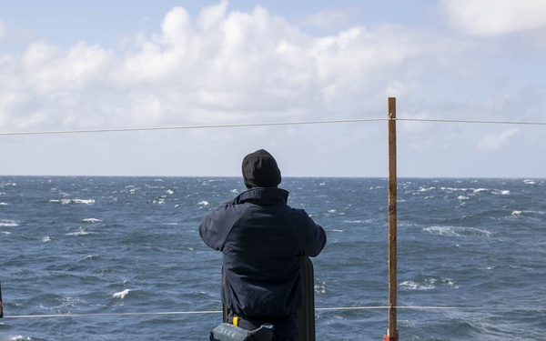 Three personnel dressed in dark navy uniforms are standing on the deck of a naval vessel at sea, setting up or adjusting black silhouette shooting targets suspended from a wire. The targets are human-shaped and appear to be part of a marksmanship or weapons training exercise. The sea conditions appear rough with choppy waves visible in the background under a partly cloudy sky.