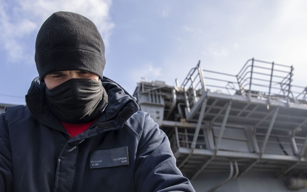 A U.S. Navy sailor dressed in cold-weather gear, including a black beanie, face covering, and insulated jacket, is performing deck work aboard a naval vessel. The sailor's name tag is partially visible and appears to read 'Thompson, USN.' The background shows the superstructure of a large gray naval ship under a partly cloudy sky.