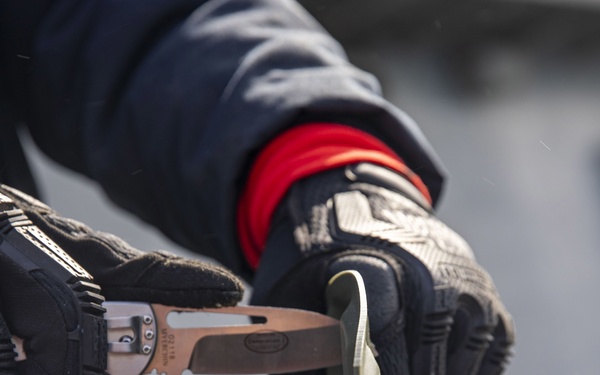A person wearing black tactical/work gloves with red wrist accents is using a folding knife or multi-tool to work on a long, flat object, possibly a blade or strip of material. The image is a close-up shot focused on the hands and tool, with a blurred background suggesting an outdoor or industrial setting. The gloves appear to be a brand like Mechanix Wear based on their distinctive styling.