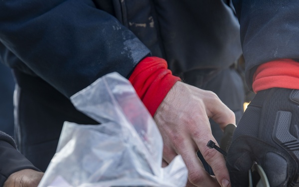 Close-up image showing hands of multiple individuals handling a clear plastic bag containing what appears to be printed documents or papers. One person is wearing a dark tactical/work glove branded 'Mechanix' with a red wrist cuff, while others have bare or lightly covered hands. The setting appears to be an outdoor or industrial environment based on the wooden surface visible in the background.