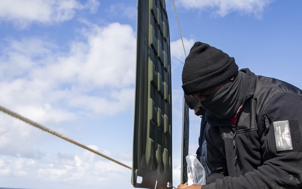 A naval personnel member dressed in all-black cold-weather gear and a face covering is working on the deck of a military vessel at sea. The sailor appears to be handling equipment or rigging near the ship's railing, with open ocean visible in the background. The ship's superstructure, including what appears to be radar or communications equipment, is visible in the upper right.