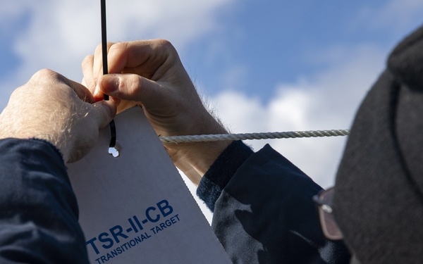 A person wearing a dark jacket and beanie is attaching or securing a cardboard shooting target to a rope or line outdoors. The target is labeled 'TSR-II-CB TRANSITIONAL TARGET' and features a human silhouette outline. The setting appears to be an outdoor shooting range under a partly cloudy sky.
