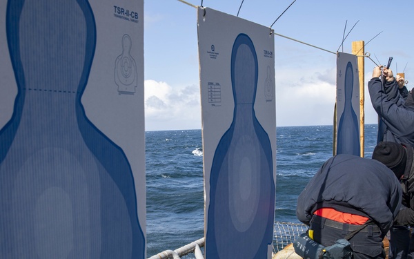 Two personnel dressed in dark tactical/naval uniforms are setting up or adjusting human-silhouette shooting targets on the deck of a naval vessel at sea. The targets are standard blue TSR-II-CB type paper silhouette targets mounted on wooden frames along the ship's railing. The setting appears to be a marksmanship training exercise conducted aboard a military ship in open ocean conditions.
