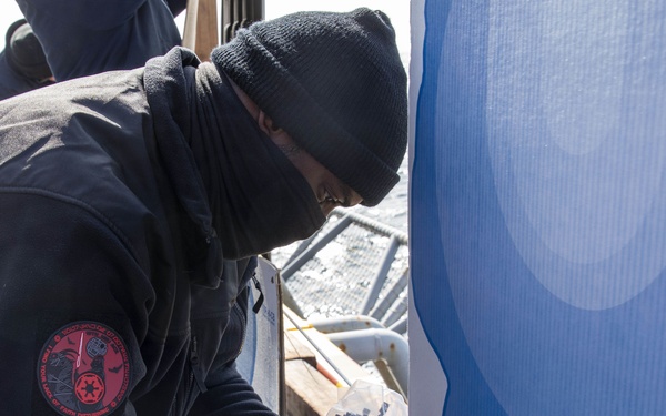 A person wearing cold-weather gear, including a black beanie and face covering, is working on attaching or securing a large blue banner or sign to a wooden frame. They are wearing a black jacket with a distinctive red circular patch on the sleeve. The setting appears to be an outdoor environment, likely on a ship or industrial platform based on the background elements.