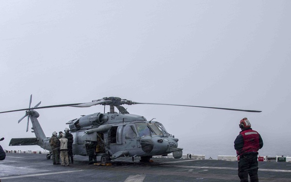 A military helicopter, appearing to be a Sikorsky MH-60 Seahawk, is parked on the flight deck of a naval vessel under overcast, foggy conditions. Several personnel in varied colored vests and gear are positioned around the aircraft and on the deck, suggesting pre-flight or post-flight operations. The scene takes place at sea, with the ocean visible in the background.