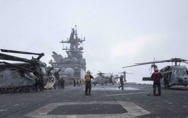 The image shows the flight deck of a U.S. Navy amphibious assault ship during adverse weather conditions, likely rain or snow. Multiple military helicopters are present on the deck, including what appear to be CH-53 Super Stallions and an MH-60 Seahawk, with flight deck crew in colored jerseys performing operations. The ship's island superstructure is visible in the background.