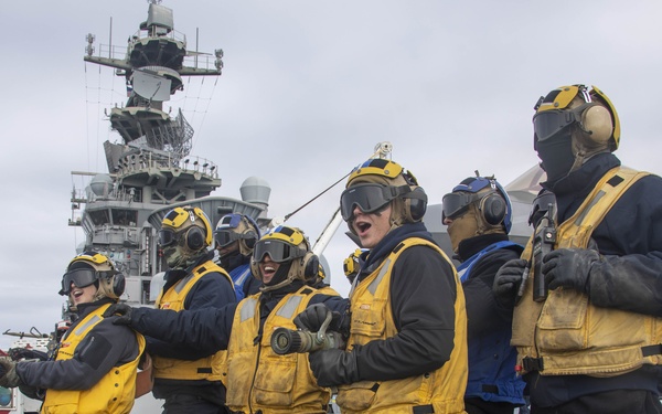 A group of naval personnel in yellow safety vests, helmets, and goggles are working on the flight deck of an aircraft carrier. They appear to be conducting a firefighting or damage control drill, holding hose equipment in a coordinated line formation. The ship's island superstructure is visible in the background under overcast skies.