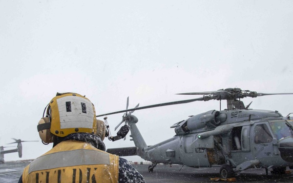 A flight deck crew member wearing a yellow 'FLT DK' vest and helmet stands on a snow-covered naval vessel flight deck, facing a gray military helicopter. The scene appears to be taking place during winter or adverse weather conditions, with snow visibly falling. Additional aircraft, including what appear to be V-22 Ospreys, are visible in the background on the left side of the deck.