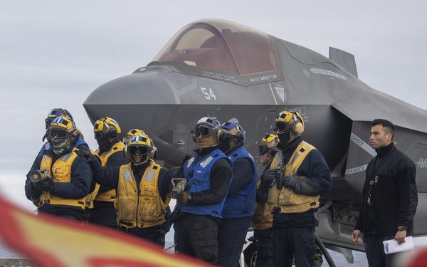 A group of naval aviation deck crew personnel stand on an aircraft carrier flight deck in front of an F-35B stealth fighter jet marked with the number '54' and squadron markings 'VMFA-122'. Crew members are wearing standard flight deck safety gear including yellow and blue vests, helmets, goggles, and hearing protection. A red and yellow flag or streamer is visible in the foreground, blurred due to depth of field.