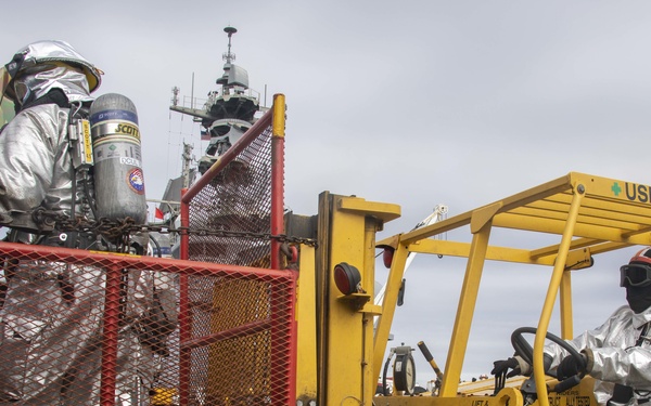 Two personnel wearing silver aluminized fire-protective suits and breathing apparatus are operating a yellow shipboard forklift (USN13-35053) on what appears to be a naval vessel deck. One operator is driving the forklift while the other rides in a red safety cage (labeled D9C154/G01Z) attached to the forks. A naval warship superstructure is visible in the background, indicating this is a pier or shipboard firefighting drill or operation.
