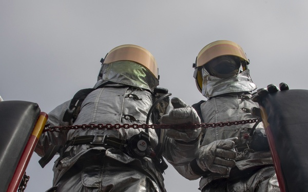 A low-angle upward shot of two figures wearing silver full-body suits and helmets with tinted visors, standing on an elevated red metal platform secured by chains. The figures appear to be statues or mannequins dressed in retro-style space or hazmat suits. The overcast sky serves as the backdrop, emphasizing the imposing scale of the figures.