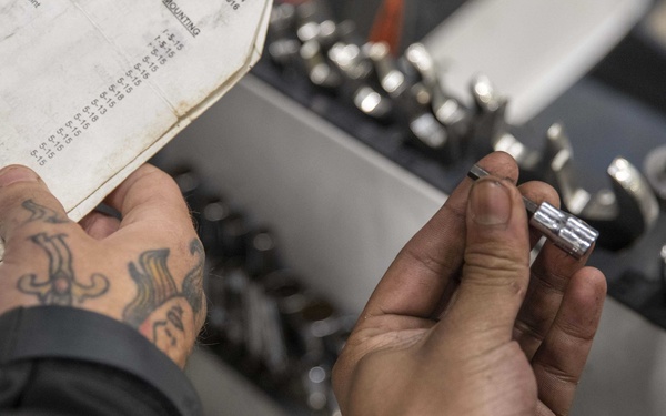 A person with visible tattoos on their left hand is examining a socket or mechanical fitting while referencing a technical document or manual. The setting appears to be a workshop or maintenance area with an organized tray of socket wrench attachments and other metal fittings. The scene suggests a mechanical repair or assembly task in progress.