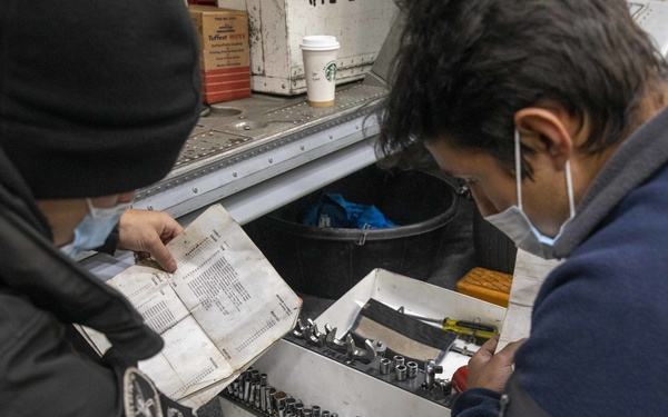 Two workers wearing face masks are crouched over an open tool chest, consulting a worn manual or reference document. They appear to be identifying or selecting specific tools, likely socket wrenches and bits, from an organized tool tray. A Starbucks coffee cup and a box of Tuffest wipes are visible in the background near what appears to be a service vehicle.