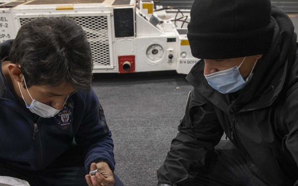 Two individuals wearing face masks are crouched on a hangar deck floor, reviewing paperwork together over a tool kit. The setting appears to be a military vessel, likely an aircraft carrier, evidenced by the fuel cart labeled 'FUEL DF-2 / JP-5' visible in the background. They appear to be conducting maintenance or inspection work using a spread of tools.