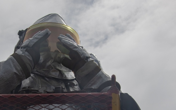 A person wearing full firefighting or hazmat protective gear, including a reflective aluminized suit and gold-visor helmet, is positioned inside a red metal mesh cage or basket, likely atop an aerial fire truck platform. The individual has their gloved hands raised to the visor of their helmet, possibly shielding their view or adjusting equipment. The photo is taken from a low angle looking upward against an overcast sky.