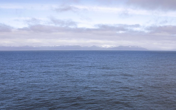A U.S. Marines MV-22 Osprey tiltrotor aircraft (bureau number 5841, tail code YT) is parked on the flight deck of a naval vessel with its rotors folded for storage. Military personnel are gathered around the aircraft on the deck. Snow-capped mountains are visible on the distant horizon across calm open water, suggesting a northern or Arctic-adjacent operating environment.