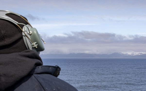 A person wearing a black hooded sweatshirt and military-style green communication headset stands on the deck of a vessel, looking out toward a calm open sea. In the distance, snow-capped mountains are visible on the horizon beneath a partly cloudy sky. The setting suggests a cold-weather maritime environment, likely a naval or coast guard operation.