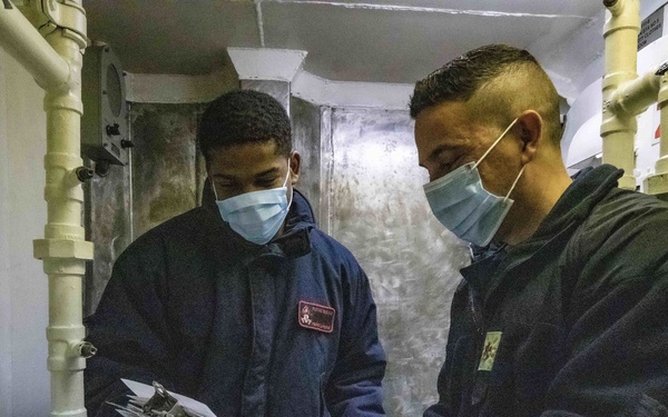Two individuals wearing blue surgical face masks and dark jackets are reviewing documents on a clipboard in what appears to be a naval vessel corridor or compartment. The space is characterized by yellow-painted pipes, white structural elements, and stenciled location markings on the wall. The setting strongly suggests a military ship interior based on the compartment labeling and infrastructure.