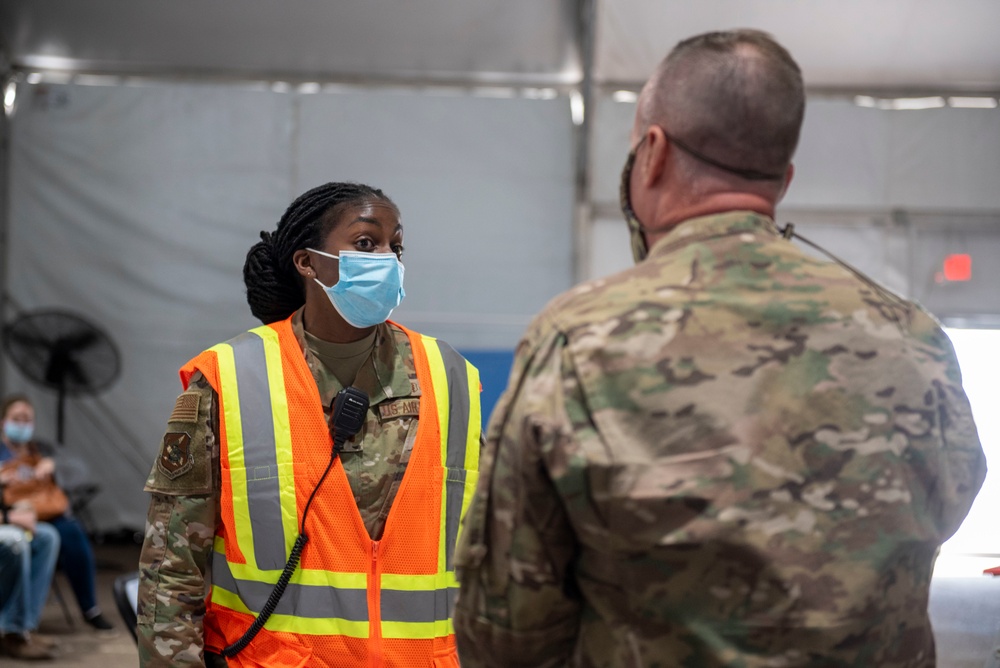 Two military personnel in camouflage uniforms are engaged in a conversation inside what appears to be a temporary tent or field facility. The soldier facing the camera is wearing an orange high-visibility safety vest, a blue surgical mask, and has a radio clipped to their vest. The setting suggests a military support operation, possibly related to COVID-19 response, disaster relief, or a similar civil assistance mission.