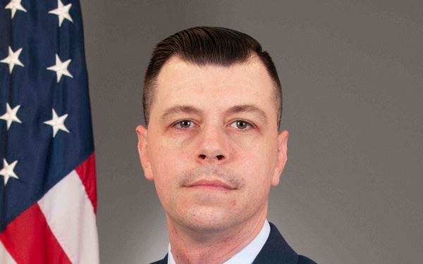 This is a formal U.S. Air Force official portrait of a male service member in dress blues uniform. He is posed in front of an American flag against a neutral gray studio background. His uniform displays a Technical Sergeant rank chevron, multiple service ribbons, occupational badges, and 'U.S.' collar insignia.
