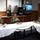 Three military personnel in U.S. Navy camouflage uniforms (NWU Type III) are seated at white-tablecloth-covered tables in what appears to be a military dining facility (DFAC/mess hall). They appear to be having a working lunch or formal dining meeting, with food plates and beverages visible on the tables. A Pepsi fountain dispenser and coffee equipment are visible in the background.